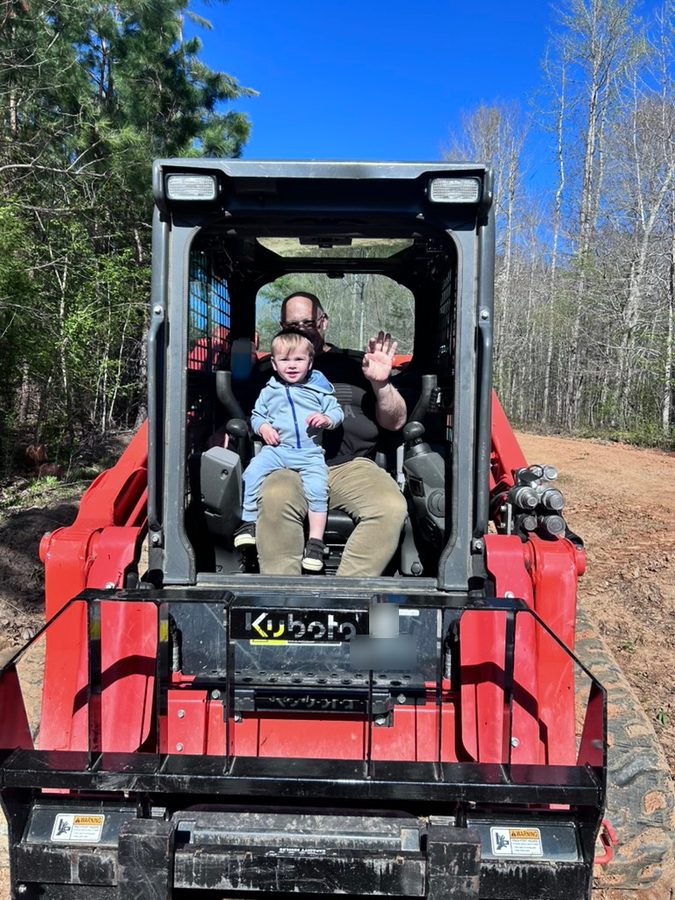 Richard French waving from the cab of his red Kubota SVL97-2 track loader with a smiling toddler on his lap, on a cleared dirt lot in the woods